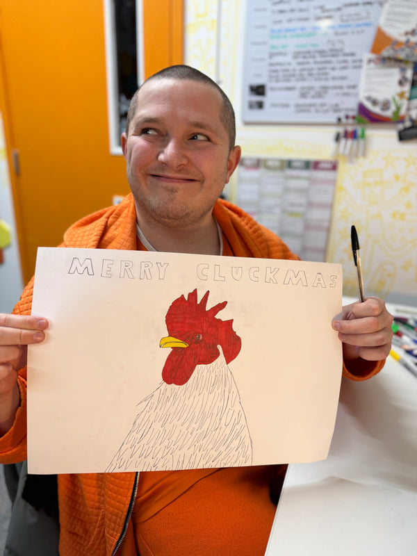 A smiling Artist holding illustration featuring a chicken with text that reads 'Merry Cluckmas'g